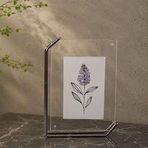Puede incluir: Un marco rectangular de acrílico transparente sostenido por un soporte de metal plateado. El marco muestra una tarjeta blanca con un dibujo lineal negro de una flor de lavanda con pétalos de color violeta claro. La estructura es vertical y sencilla.