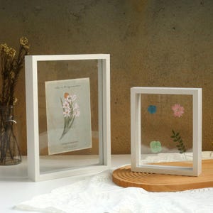 Puede incluir: Dos marcos de fotos blancos con frentes de vidrio. El marco más grande tiene una flor prensada en su interior. El marco más pequeño tiene flores y hojas prensadas en su interior.