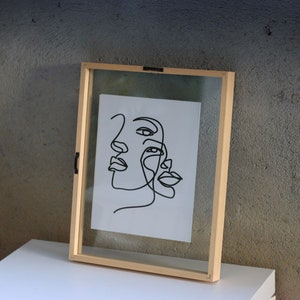 May include: A rectangular floating picture frame with a light-colored wooden border containing a white sheet of paper. The paper features a black continuous line drawing of two overlapping human faces, showcasing eyes, noses, and lips in profile.