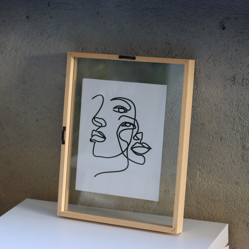 May include: A rectangular floating picture frame with a light-colored wooden border containing a white sheet of paper. The paper features a black continuous line drawing of two overlapping human faces, showcasing eyes, noses, and lips in profile.