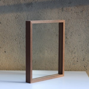 May include: A rectangular floating frame with a medium-brown wood grain finish. The frame features a deep profile and a clear glass center, designed to hold items between two panes of glass, creating a transparent display effect for artwork.