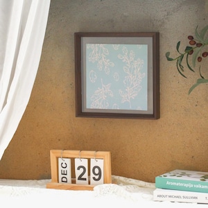 Puede incluir: Un marco con una impresión con fondo azul claro y diseño floral blanco cuelga de una pared texturizada. Un calendario de madera muestra "DIC 29". Un libro con texto verde y blanco es visible.