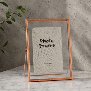 Puede incluir: Un marco de fotos rectangular de metal en color oro rosa con un borde de cuentas. El marco cuenta con dos paneles de vidrio transparente que sostienen un inserto blanco con el texto "Photo Frame" y "GENTLETIMES 4x6". Se apoya en un soporte fino.