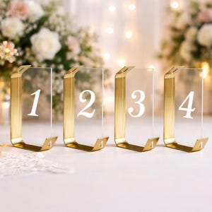 Puede incluir: Cuatro letreros rectangulares de acrílico transparente para mesas, cada uno sostenido por un soporte de metal dorado. Los letreros están numerados del uno al cuatro con una fuente serif blanca. Cada soporte de metal tiene un pequeño recorte triangular.