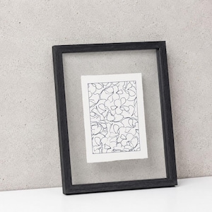 May include: A rectangular floating picture frame with a black wooden border containing a white paper insert. The insert features a black ink line drawing of overlapping floral shapes, creating a continuous pattern across the rectangular surface.