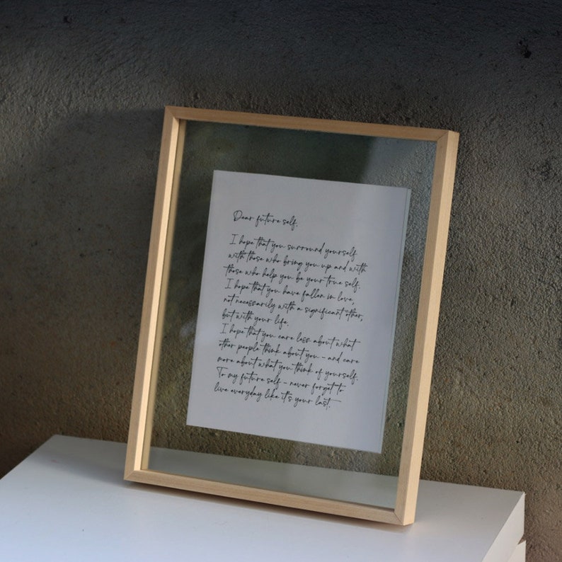 May include: A rectangular light wood frame containing a white paper with black cursive text that reads: Dear future self. I hope that you surround yourself with those who bring you up and with those who help you be your true self. I hope that you have fallen in love, not necessarily with a significant other, but with your life. I hope that you care less about what other people think about you - and care more about what you think of yourself. To my future self - never forget to live everyday like it's your last.