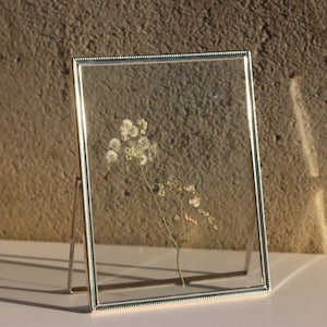 Könnte beinhalten: Ein silberner Bilderrahmen mit einer Perlenbordüre, der auf einer weißen Oberfläche steht. Der Rahmen enthält eine getrocknete Blumenarrangements mit zarten weißen Blüten.