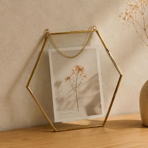 May include: A hexagonal gold metal frame with clear glass panels, suspended by a delicate gold chain. Inside the frame is a rectangular print featuring a single botanical stem with small flowers and the text As long as the sun flowers will bloom.