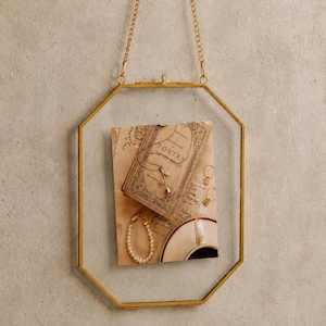 May include: A hanging octagonal picture frame with a thin gold metal border and a matching gold chain. Inside the clear glass panes is a rectangular print featuring a vintage book titled Poètes du XIXe Siècle, a pearl bracelet, and jewelry.