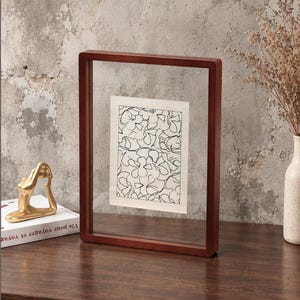 May include: A rectangular floating picture frame with a dark brown wooden border and two clear glass panes. Inside the frame is a white rectangular paper insert featuring a black line drawing of overlapping floral shapes and petals.