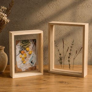 Può includere: Due cornici rettangolari in legno chiaro con design flottante. La cornice più piccola contiene una fotografia di limoni su un tessuto, mentre quella più grande racchiude steli botanici essiccati pressati tra due lastre di vetro trasparente.