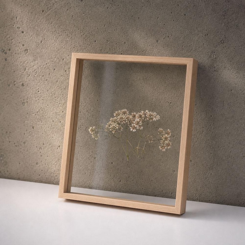 May include: A rectangular light-colored wooden floating frame containing a single sprig of dried white baby's breath flowers pressed between two clear glass panes. The frame has a simple, clean-lined construction and a smooth, natural wood finish.