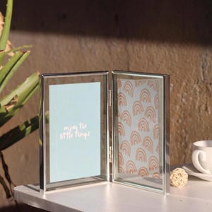 May include: A double-sided silver metal picture frame with glass panels. The left side contains a light blue insert with the white text "enjoy the little things," while the right side features a pattern of repeating orange rainbows.