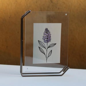May include: A clear acrylic picture frame with a metal stand. The frame holds a simple line drawing of a purple flower.