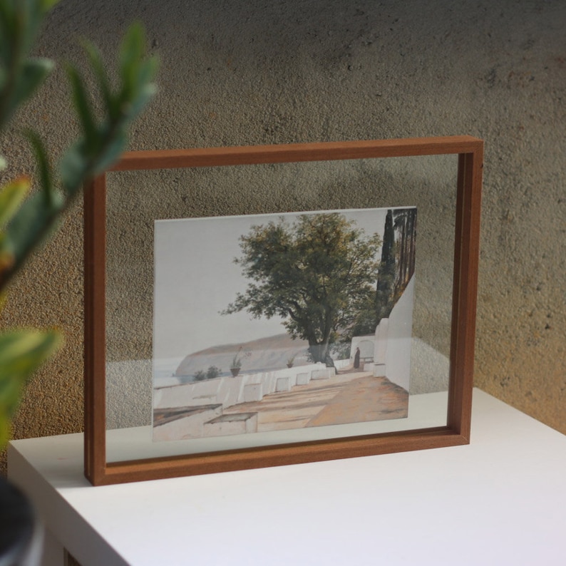 May include: A rectangular wooden floating frame containing a landscape print. The artwork depicts a sunlit coastal path with white walls, a large green tree, and a distant cliffside overlooking the sea. The frame has a natural brown wood finish.
