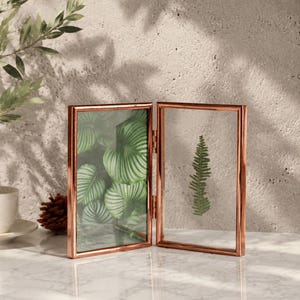 May include: A hinged double picture frame with a metallic copper finish and clear glass panes. The left frame contains a green leaf pattern, while the right frame holds a single pressed green fern leaf. The frame is rectangular and stands upright.