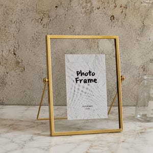 Puede incluir: Un marco de fotos de metal dorado con forma rectangular y soporte pivotante. El marco contiene una inserción blanca con un patrón de hojas dibujado en líneas negras y el texto "Photo Frame" y "GENTLETIMES 4x6" impreso en el centro.