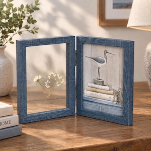 Puede incluir: Un marco de fotos doble articulado con acabado de veta de madera azul. El lado izquierdo tiene un panel de vidrio transparente con una flor blanca prensada. El lado derecho contiene una ilustración de un pájaro sobre libros con el texto Cote d'Azur.