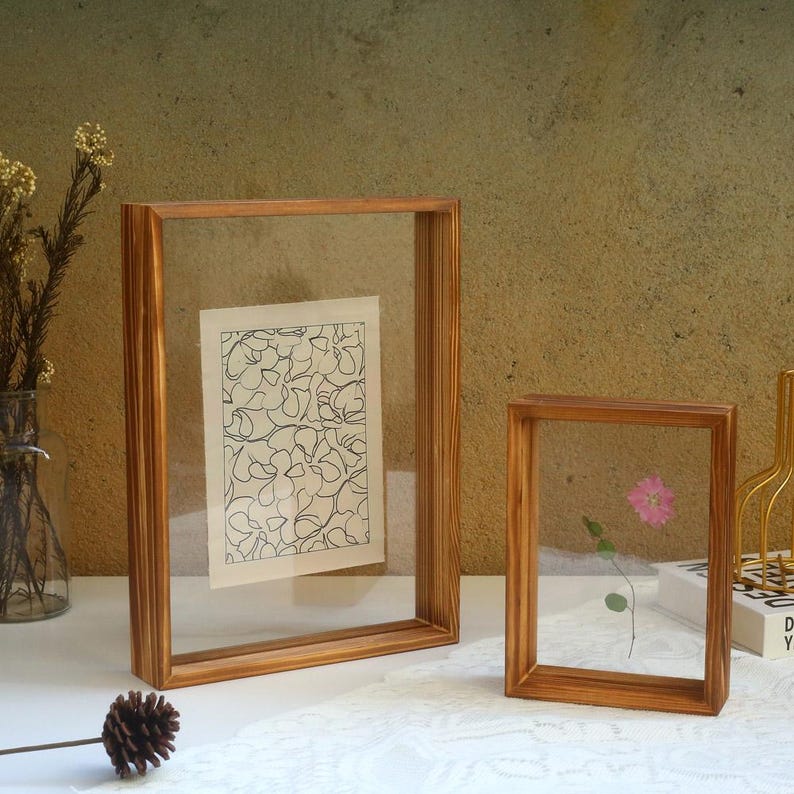May include: Two wooden picture frames with clear glass fronts. The larger frame is holding a black and white line drawing of a floral pattern. The smaller frame is holding a pressed flower.