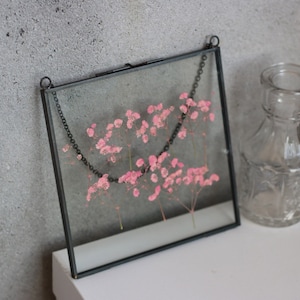 May include: A black metal frame with a glass front, holding a pressed flower arrangement of pink baby's breath. The frame is hanging from a black chain.