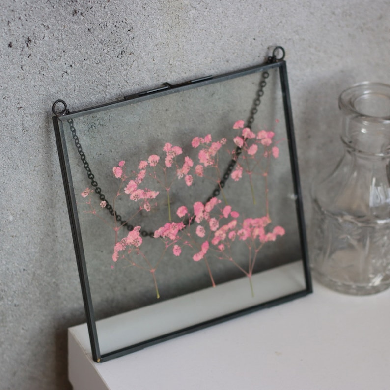 May include: A black metal frame with a glass front, holding a pressed flower arrangement of pink baby's breath. The frame is hanging from a black chain.