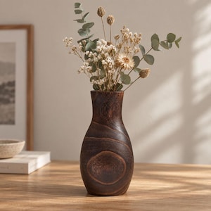 Op de afbeelding: Een donkerbruine houten vaas met een glad, rond lichaam en een smalle hals, gevuld met een boeket droogbloemen waaronder eucalyptusstengels, kleine witte gipskruidbloemen en een enkele lichtgekleurde bloem die lijkt op een madeliefje.