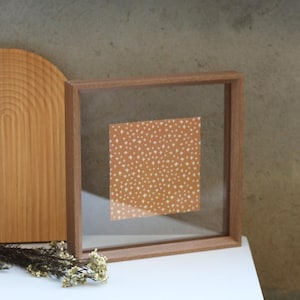 Puede incluir: Un marco de fotos cuadrado de madera con una parte frontal de vidrio transparente. Dentro del marco hay una impresión cuadrada con un fondo marrón y un patrón de pequeñas formas de estrellas blancas. Una tabla de cortar de madera y flores secas están en el fondo.