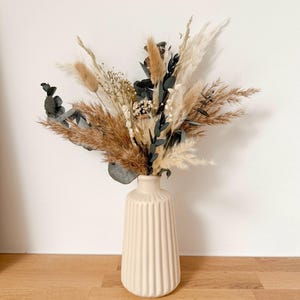 Op de afbeelding: Een crèmekleurige vaas met verticale ribbels bevat een droogbloemarrangement. Het arrangement bevat beige pampasgras, tarwe en donkergroene eucalyptusbladeren. De tekst "Variante 1" staat onderaan.