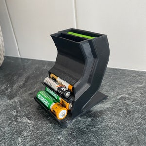 May include: A black 3D-printed battery organiser holding AA and AAA batteries. The curved design has compartments for different battery sizes. Some batteries are labelled "Rechargeable".