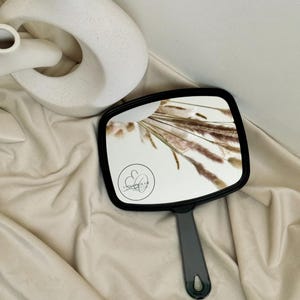 May include: A black hand mirror with a square frame and a black handle. The mirror reflects dried beige and brown grasses.