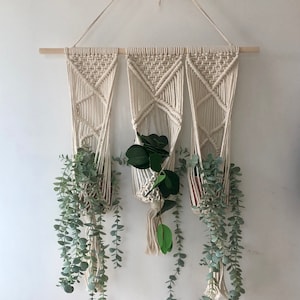 May include: Three macrame plant hangers with a wooden dowel rod, hanging on a white wall. Each hanger has a potted plant with trailing greenery.