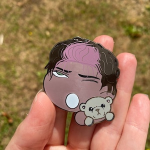 May include: A black and brown enamel pin featuring a cartoon character with pink hair and a winking expression. The character is holding a small brown teddy bear.