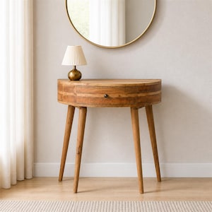 May include: A semi-circular wooden console table with a single drawer featuring a small dark knob, supported by four tapered legs. A small table lamp with a pleated white shade and a gold base sits on the left side of the tabletop surface.