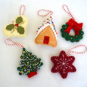 May include: A collection of handmade Christmas ornaments. The ornaments include a holly-shaped ornament, a gingerbread house, a wreath with a red bow, a Christmas tree, and a red star with a snowflake design. Each ornament has a red and white string for hanging.