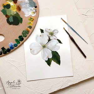 May include: A watercolour painting of white dogwood blossoms with green leaves. The painting is on a white background and is surrounded by a wooden palette and paintbrushes.