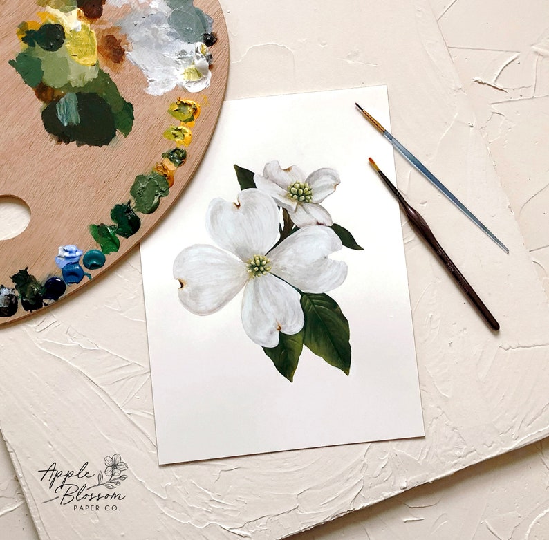 Puede incluir: Una pintura de acuarela de flores blancas de cornejo con hojas verdes. La pintura est&aacute; sobre un fondo blanco y est&aacute; rodeada de una paleta de madera y pinceles.