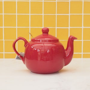 May include: A red ceramic teapot with a shiny finish. The teapot has a handle and a spout. It is sitting on a white surface.