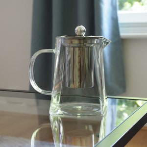 May include: A clear glass teapot with a stainless steel lid and strainer. The teapot has a handle and a spout. The teapot is sitting on a glass table.
