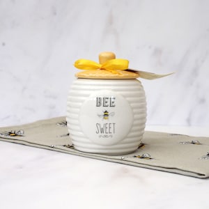 May include: White ceramic honey pot with a yellow lid and a yellow ribbon. The pot has a bee design and the text "BEE SWEET".