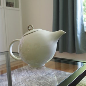May include: A white ceramic teapot with a silver lid and handle. The teapot is sitting on a glass table with a white rug underneath.