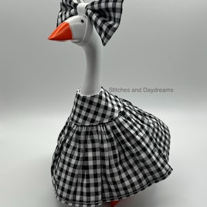 May include: A white plastic goose figurine wearing a black and white gingham dress and a matching bow on its head. The orange beak is visible. The text Stitches and Daydreams is printed on the right side of the goose. The figure has a smooth finish.