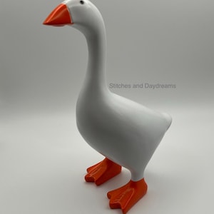 May include: A white goose figurine with a bright orange beak and orange webbed feet. The goose is standing in a profile position, facing left. The text Stitches and Daydreams is printed in a light gray font on the body of the goose figurine.