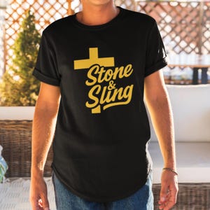 May include: Black t-shirt with a gold cross graphic and the words "Stone & Sling" in a stylized font. The shirt has rolled-up sleeves and a crew neck. The design is centered on the chest.