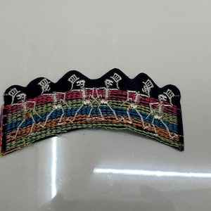 Puede incluir: Parche bordado con una fila de esqueletos bailando en hilo blanco. Los esqueletos se colocan sobre un fondo de arcoíris y un borde negro festoneado. Un accesorio único y colorido.