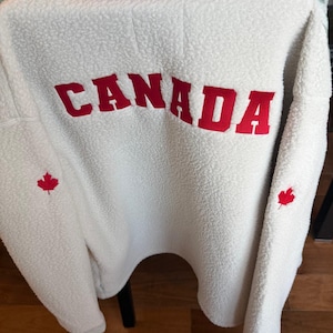 Può includere: Un maglione in pile sherpa bianco con la scritta CANADA in grandi lettere rosse sul retro. Una sottile striscia orizzontale rossa si trova sopra il testo e un piccolo emblema a forma di foglia d'acero rossa è ricamato su ciascuna manica.