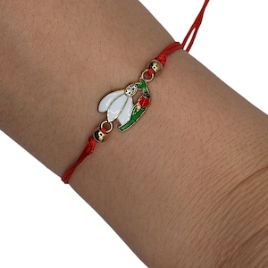 Op de afbeelding: Rood koord armband met een gouden bedel met een witte sneeuwklokje bloem met groene bladeren en een rode lieveheersbeestje.