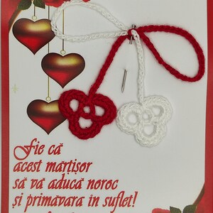 May include: A red and white crocheted martisor with a message that reads "Fie ca acest martisor sa va aduca noroc si primavara in suflet! Martie". The martisor is on a white card with red roses and hearts.