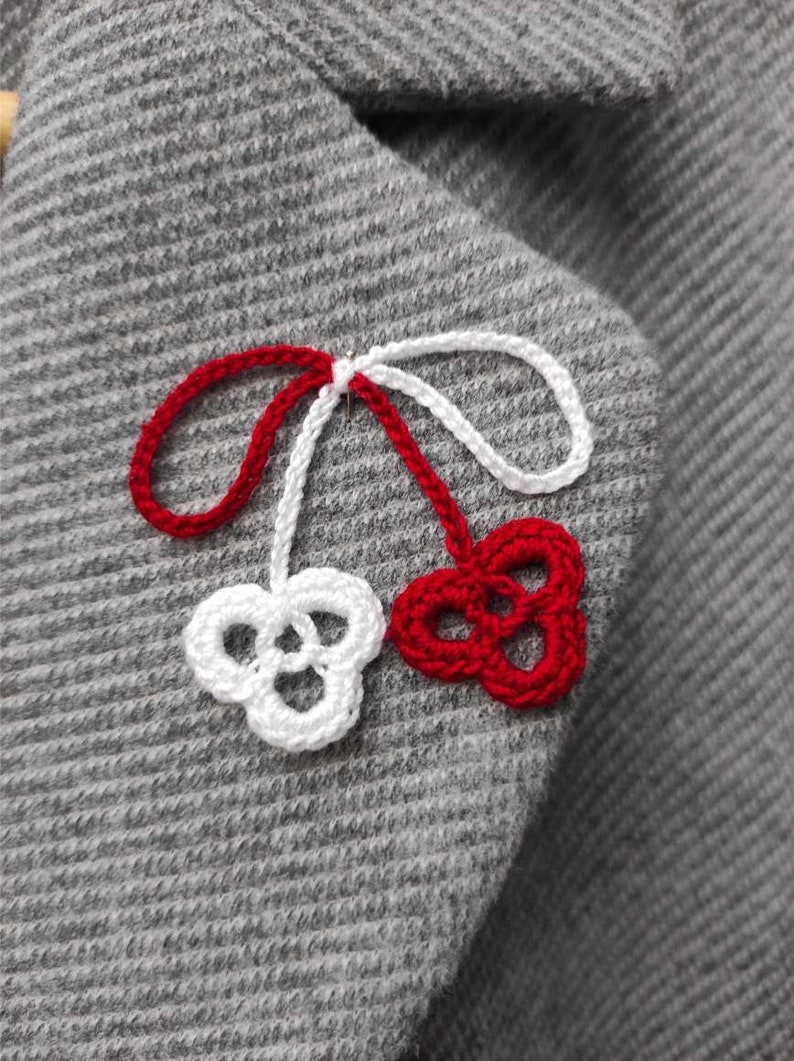 May include: A close-up of a gray knitted fabric with a red and white crocheted cherry design. The cherry has a stem with a loop at the top.