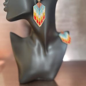 May include: A pair of long, colourful beaded earrings. The earrings are made of small, colourful beads that are woven together in a geometric pattern. The earrings are a vibrant mix of red, orange, yellow, blue, and white beads.