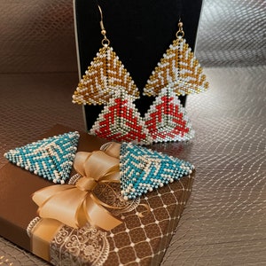 Puede incluir: Un par de pendientes de forma triangular con patrones geométricos en dorado, blanco, rojo y azul. Los pendientes se muestran en una pequeña caja de regalo con un diseño de patrón marrón y blanco.